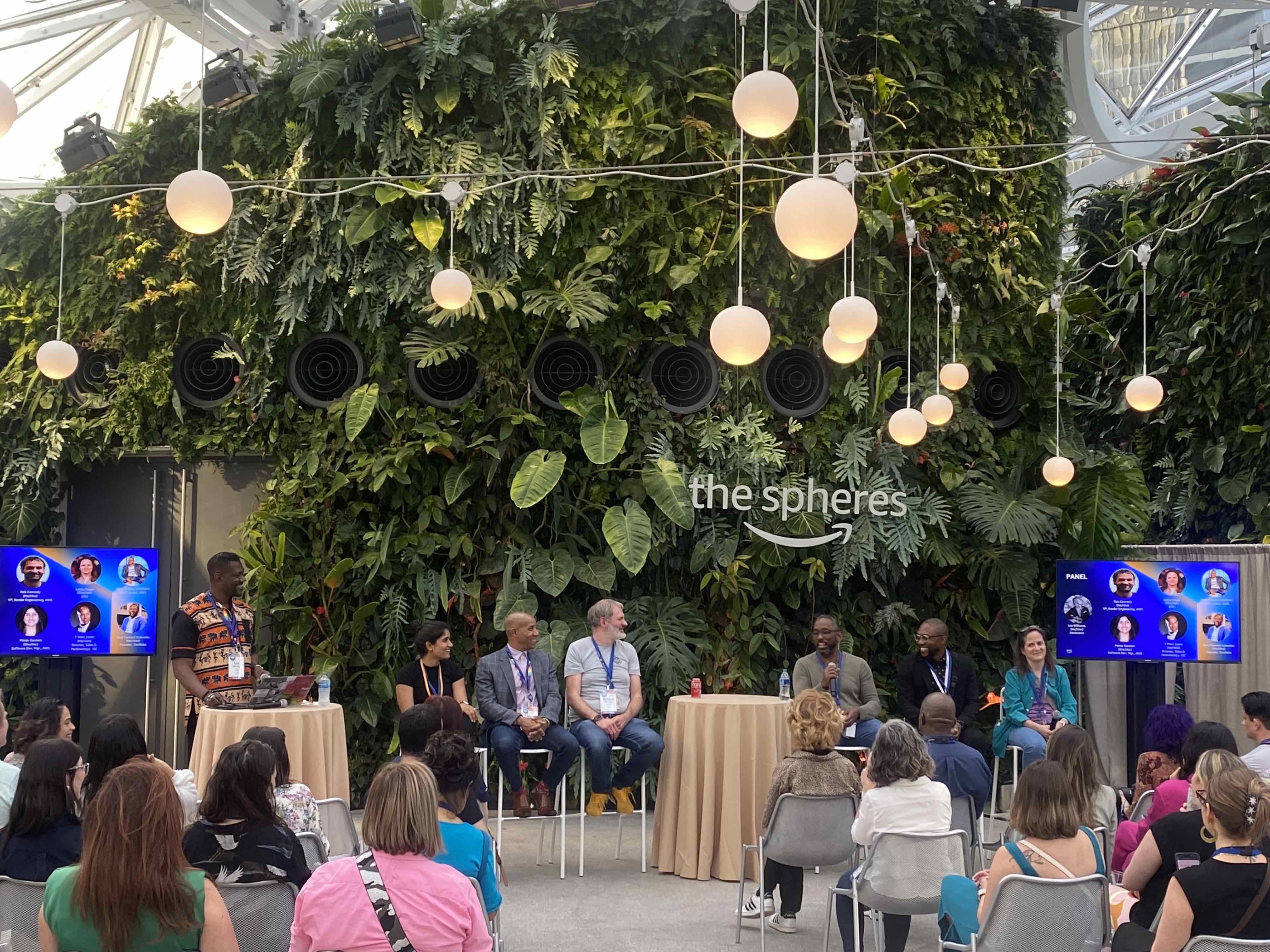 Six panelists on stools with a multi-storey backdrop wall of lush plants